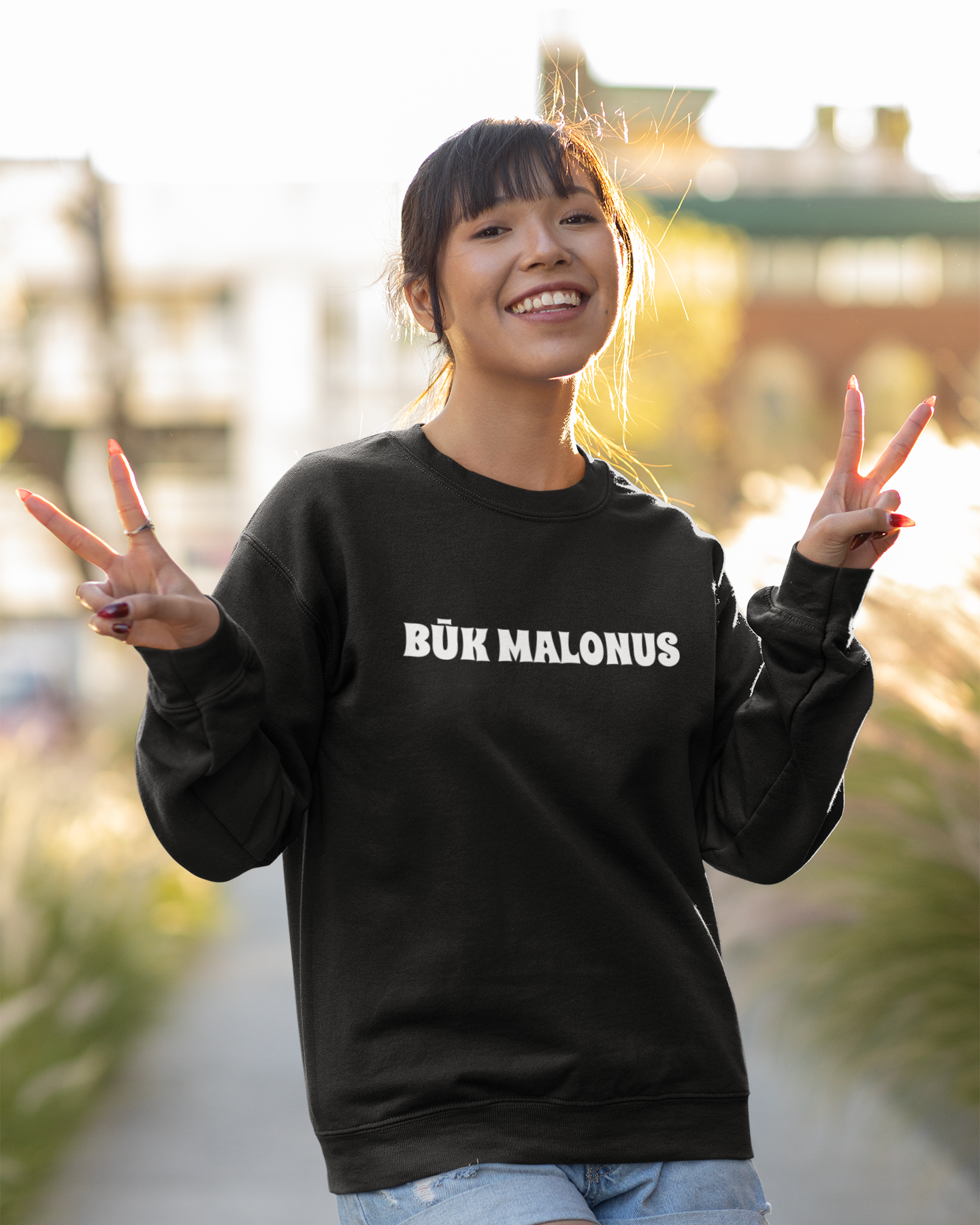 Pozuojanti moteris parke, rodanti taikos gestus, vilki juodą lietuviškos tematikos džemperį su užrašu "BŪK MALONUS".