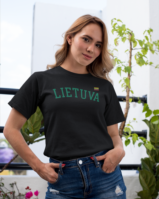 Moteris, stovinti prie augalų su juodais marškinėliais, puoštais užrašu „LIETUVA“ ir nedidele Lietuvos trispalve prie širdies