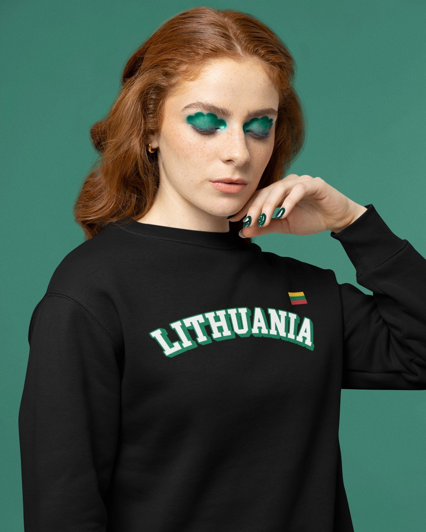 Rausvaplaukė moteris su ryškiu žaliu makiažu dėvi juodą džemperį su užrašu „LITHUANIA“ ir Lietuvos trispalve širdies plote
