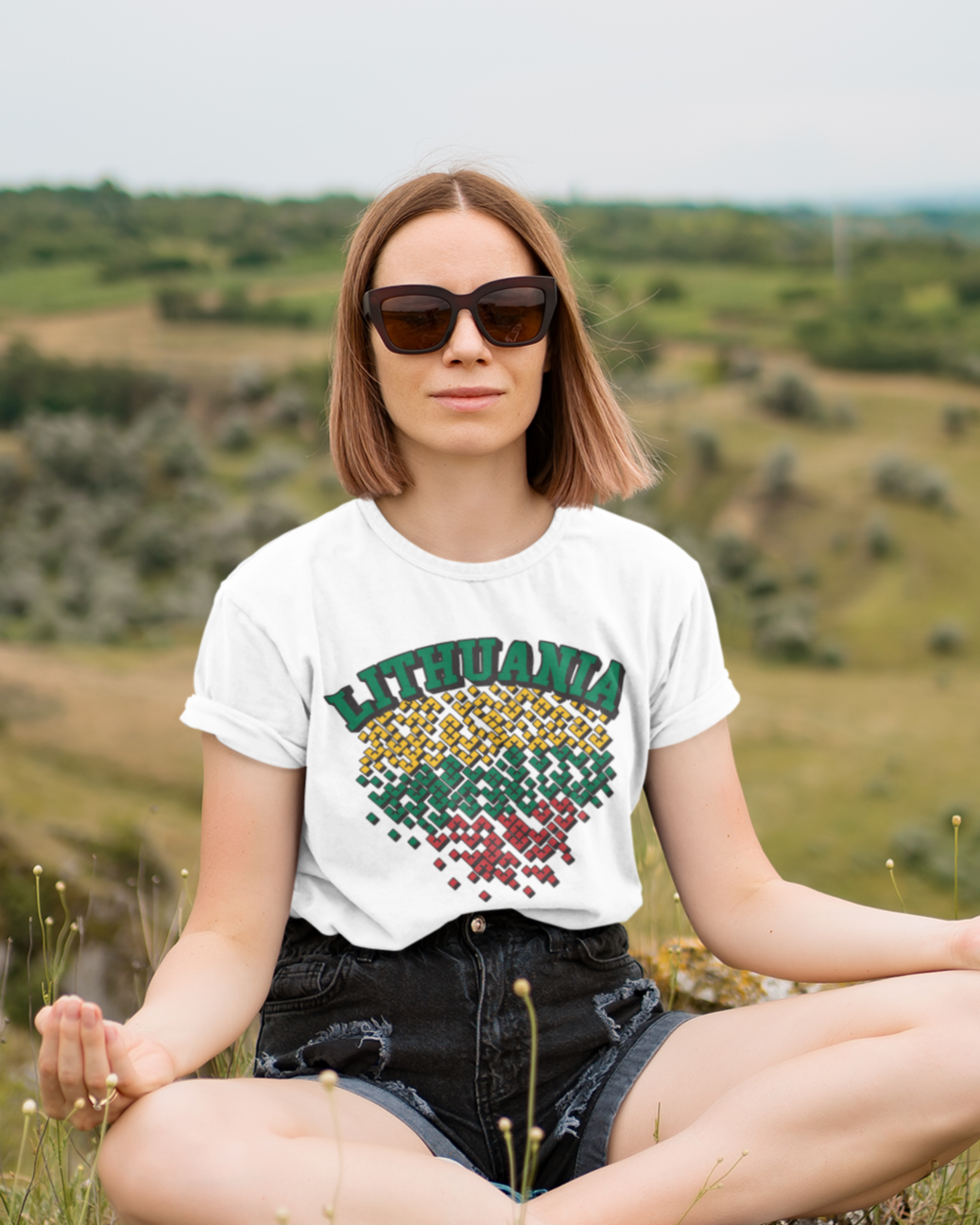 Gamtoje sėdinti moteris dėvi baltus marškinėlius su užrašu „LITHUANIA“ ir trispalvės spalvomis nuspalvintas tautines detales