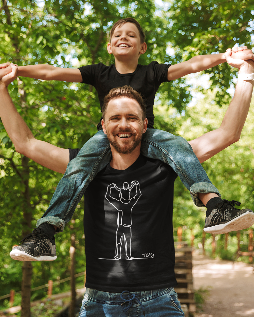 Linksmas ir emocingas kadras, kai tėvas su sūnumi ant pečių dėvi juodus marškinėlius su linijiniu piešiniu ir užrašu „Tėtis“.