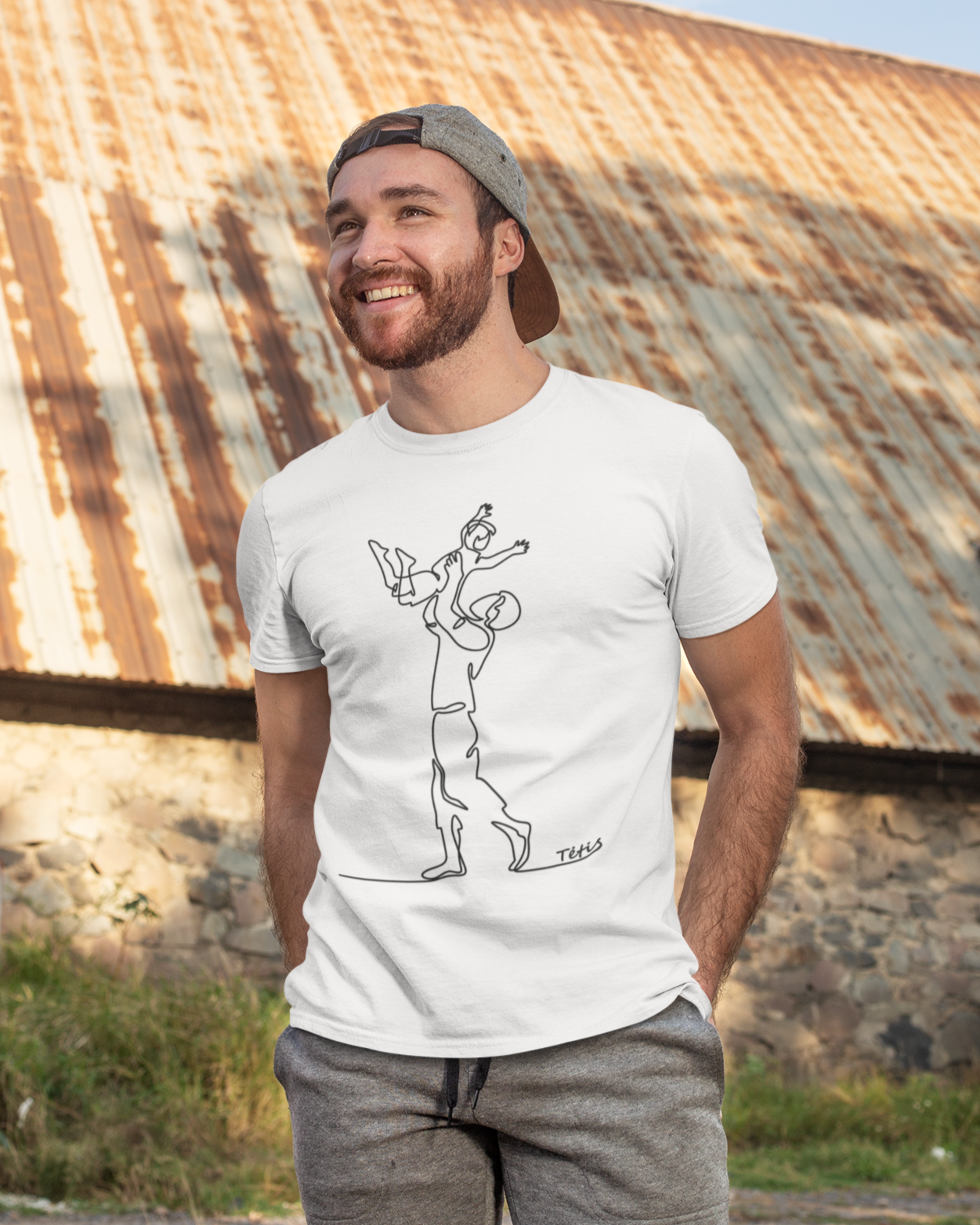 Barzdotas vyras lauke apsivilkęs kepure ir kelnėmis, priderinęs baltą maikutę su tėčio dienos atributikos linijiniu piešiniu.