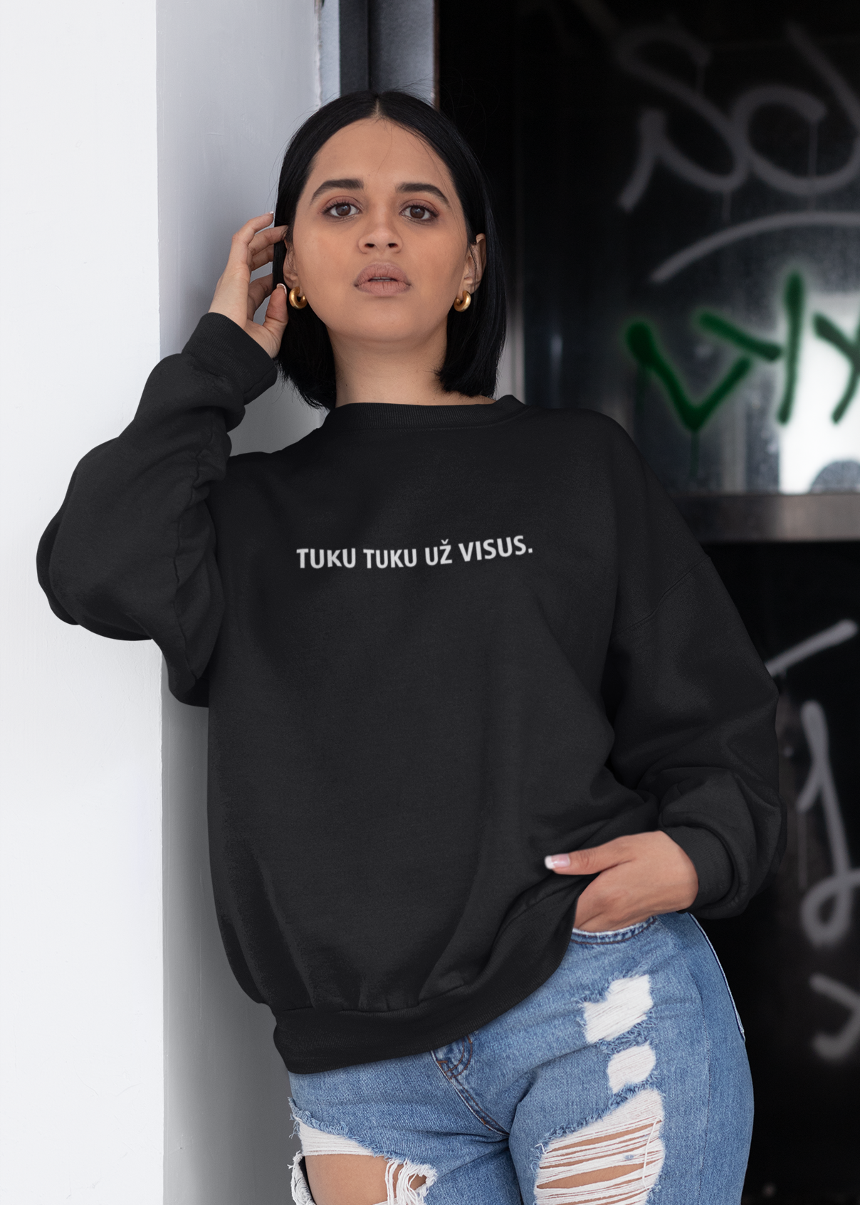 Atsirėmusi į sieną, miesto stiliaus kadre, jauna moteris dėvi juodą džemperį su užrašu „TUKU TUKU UŽ VISUS.“