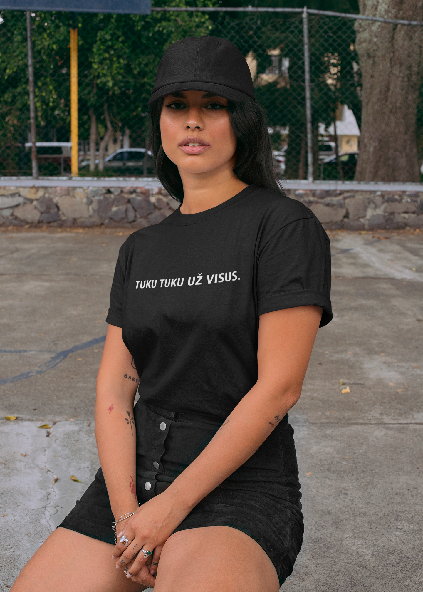 Jauna moteris sėdi lauko sporto aikštelėje dėvėdama juodus marškinėlius su baltu užrašu „TUKU TUKU UŽ VISUS“.