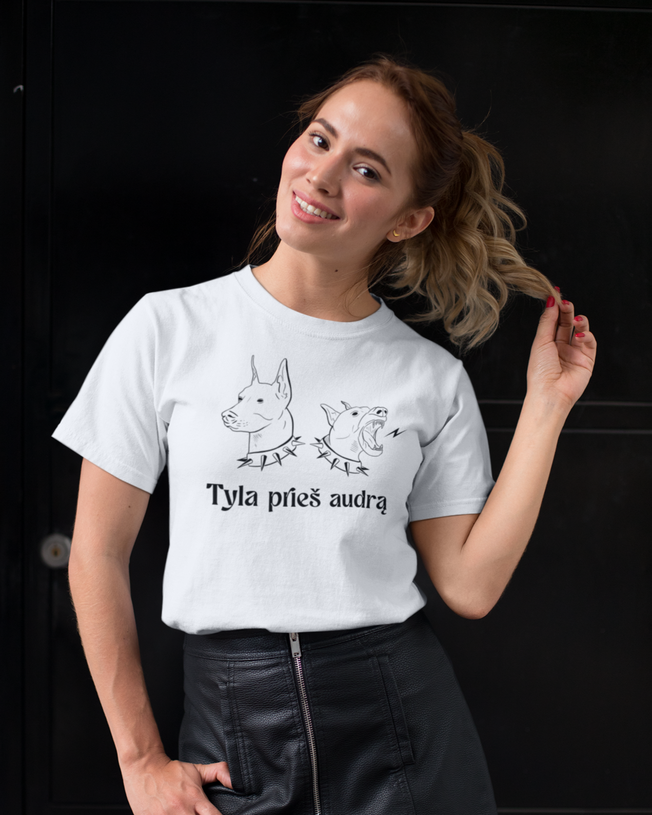Moteris šypsosi, vilkėdama baltus lietuviškus marškinėlius su dviem šunų galvų iliustracijomis ir užrašu „Tyla prieš audrą“.