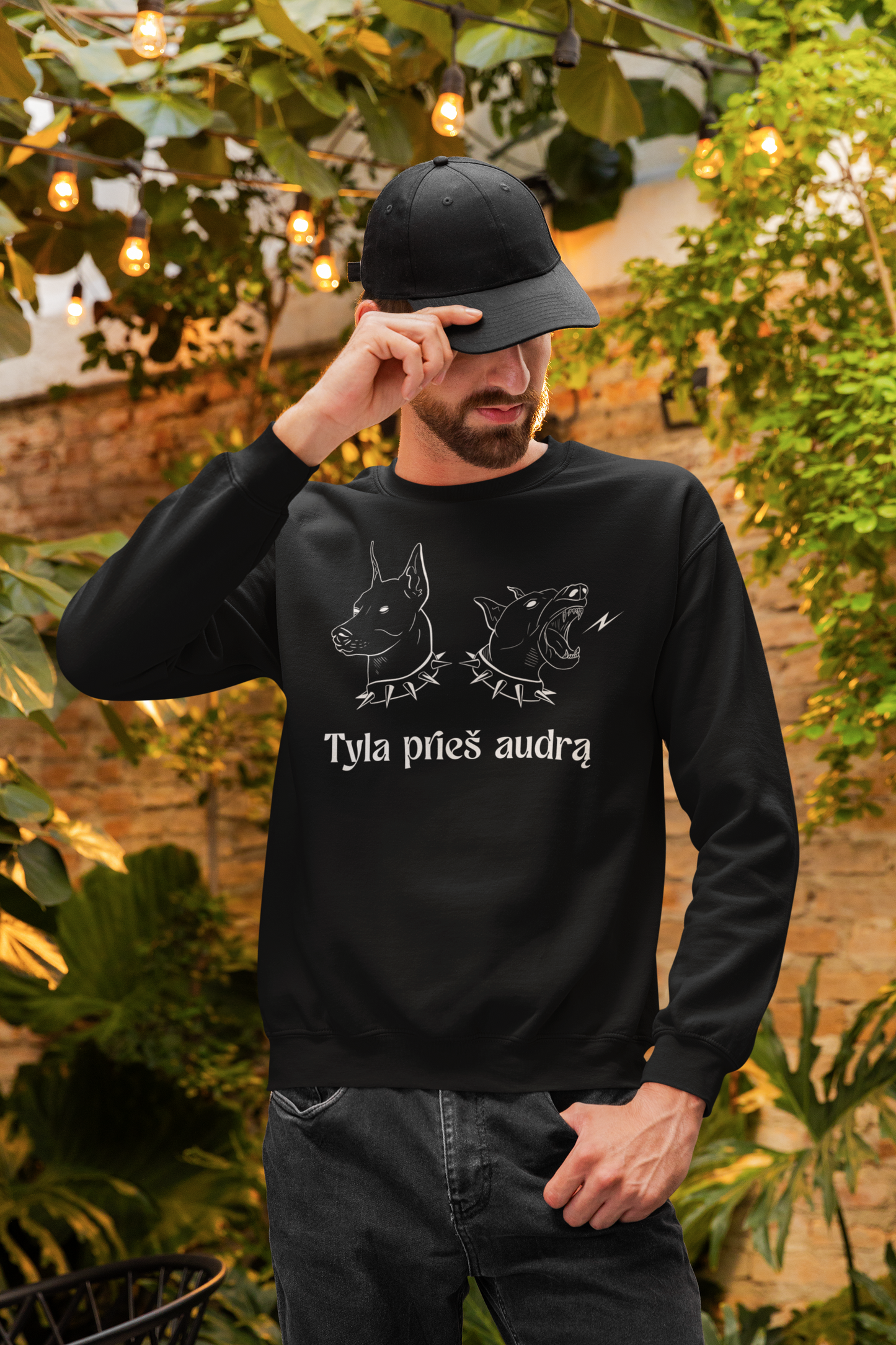 Vyras su lietuvišku juodu džemperiu „Tyla prieš audrą“ stovi lauke, tarp augalų ir lempučių, laikydamas kepurės snapelį.