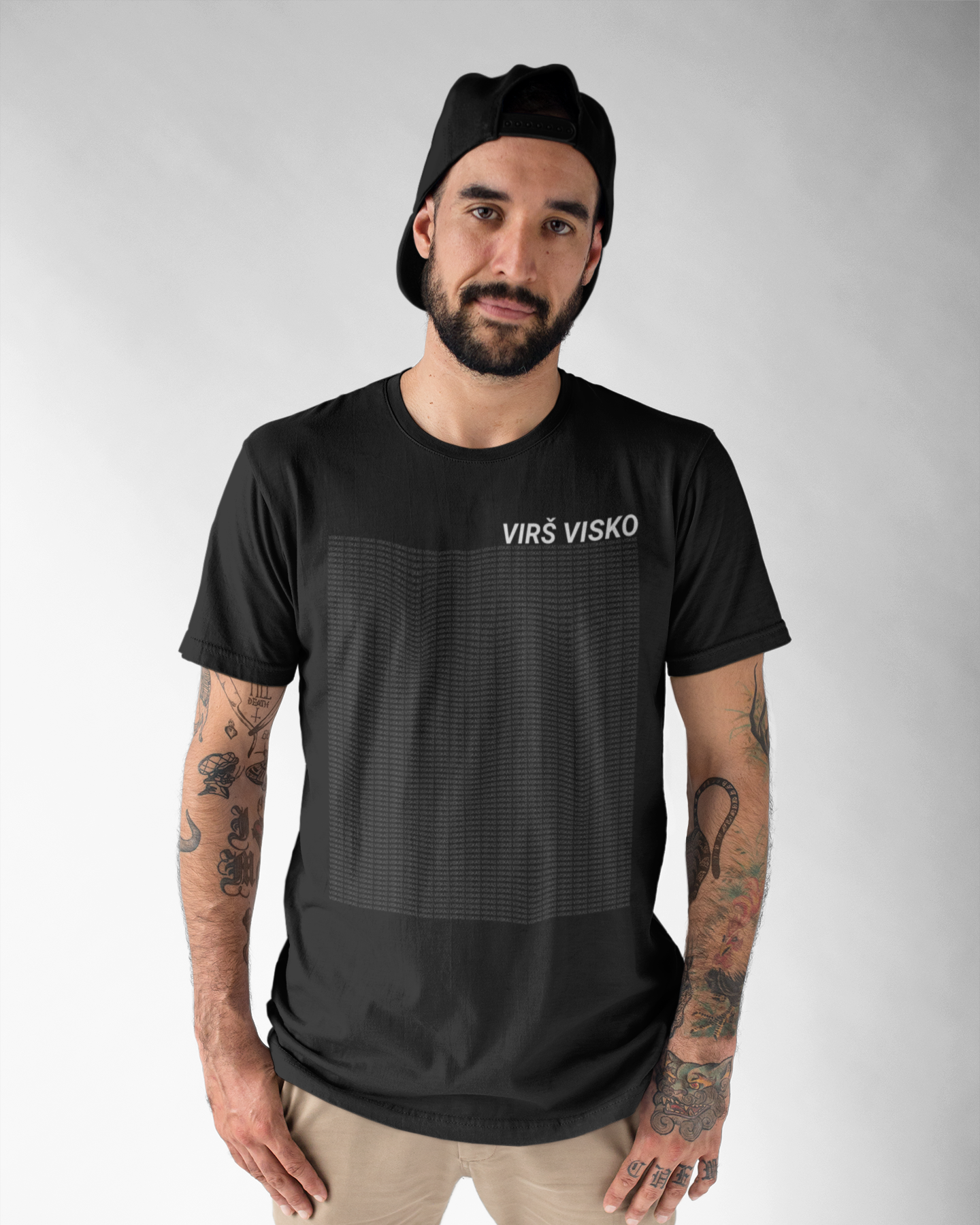 Vyras prie smėlio spalvos kelnių ir kepuraitės priderinęs juodus marškinėlius su kontrastingu baltu užrašu „VIRŠ VISKO“.