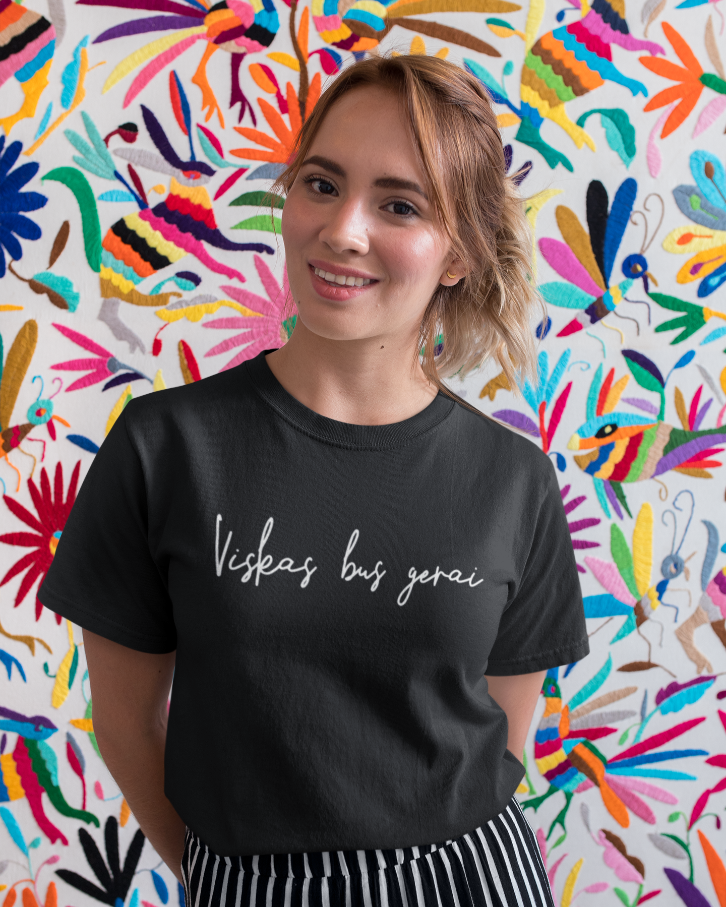 Besišypsanti mergina, spalvingame fone su juodais marškinėliais su minimalistiniu užrašu lietuviškai „Viskas bus gerai“.