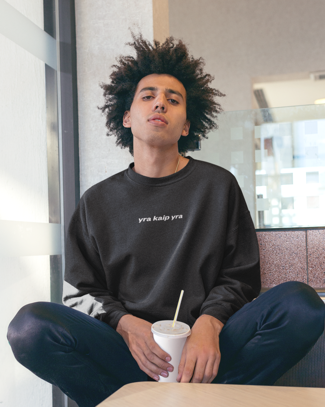 Vyras sėdintis kavinėje dėvi juodą džemperį su lietuvišku posakiu „yra kaip yra“.