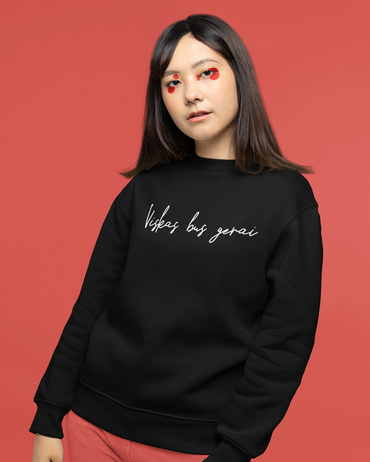 Jauna mergina apsivilkusi juodą džemperį su ranka rašytu minimalistiniu lietuviško dizaino užrašu „Viskas bus gerai“.